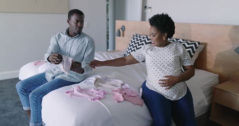 African american expectant couple sorting pink baby clothes on bed preparing nursery