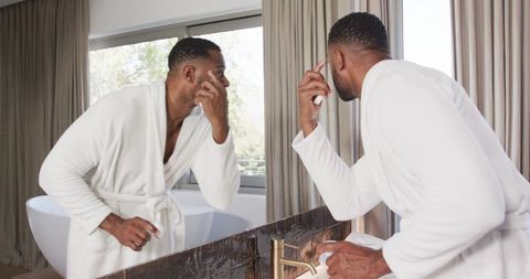 Man in white robe applying facial cream at bathroom mirror, morning grooming routine