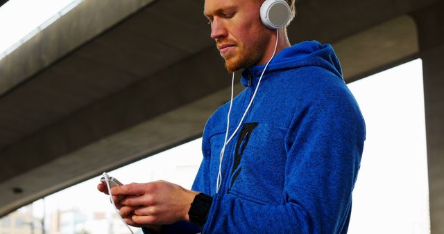 A man in a blue hoodie is under a large bridge. He looks at his smartphone while wearing headphones. Use this image for themes like outdoor activities, leisure, technology, or fitness. Suitable for promoting music streaming services, health and fitness apps, or urban lifestyle brands.
