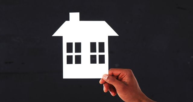 A hand holds a paper cutout of a house against a dark background, with copy space. It symbolizes concepts of home ownership, real estate investment, or the idea of finding a home.