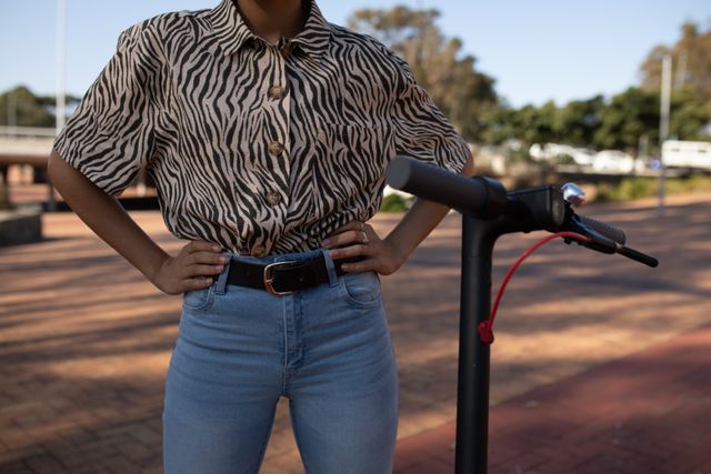 Front view mid section of a biracial woman enjoying free time in a city on a sunny day, standing with hands on hips in an urban park next to an electric scooter, wearing blue jeans and zebra print shirt.