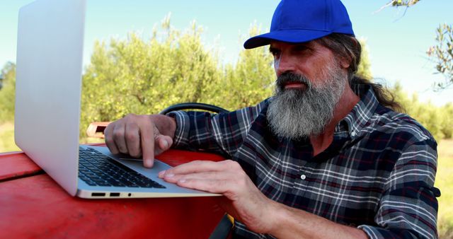 A middle-aged Caucasian man with a beard works on a laptop outdoors, with copy space. His casual attire and the rural setting suggest he might be managing agricultural tasks or enjoying leisure time in nature.