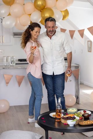 Mature couple toasting with rosé at home celebration with charcuterie board, balloons, pastel decor