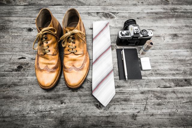 Rustic composition of vintage leather shoes, striped tie, old camera, and small accessories on wooden surface. Ideal for themes related to vintage styling, photography, accessories arrangement, and fashion trends. Can be used for blog posts, advertisements, and social media content focusing on retro and vintage appeal.