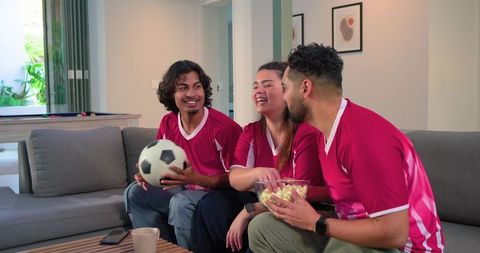 Diverse friends watching soccer at home laughing, sharing popcorn, holding soccer ball