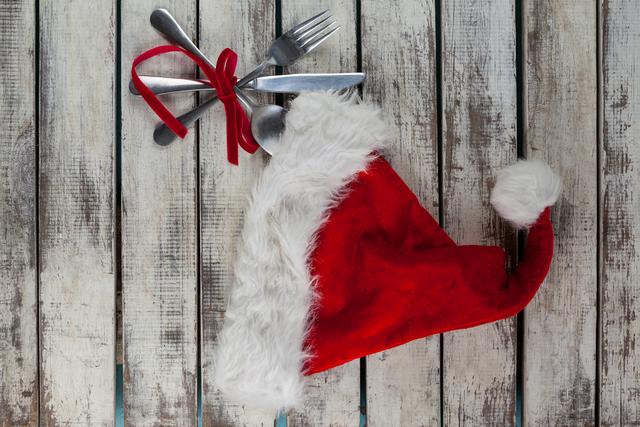 This image shows a set of cutlery tied with a red ribbon next to a Santa hat on a rustic wooden background. Ideal for holiday-themed promotions, festive dining advertisements, Christmas event invitations, and seasonal blog posts.