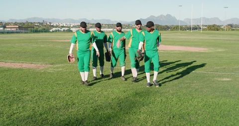 Five baseball teammates walking across sunlit outfield wearing green uniforms and carrying gloves