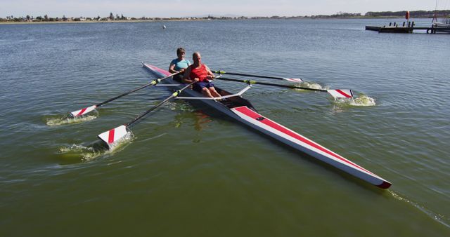 Two men rowing together in a double scull on calm lake water under a clear sky. Ideal for showcasing teamwork, outdoor activities, healthy lifestyles, or promotional material for rowing clubs.