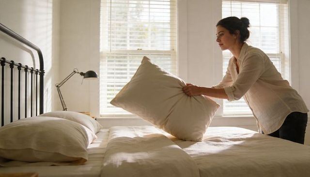 Sunlit woman making bed in minimalist neutral bedroom morning adjusting pillow and linen