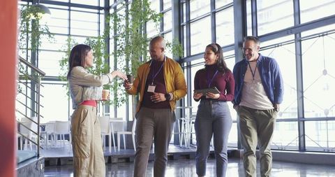 Diverse team walking and networking in modern glass atrium holding coffee and tablet