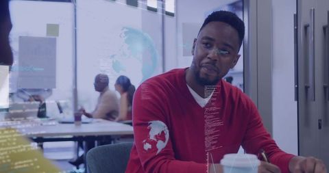 Man wearing red sweater taking notes during collaborative tech meeting in open-plan office