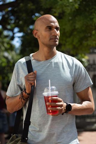 Bald man standing in sunlit park holding red iced drink wearing shoulder bag and smartwatch