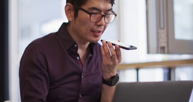 Asian professional man speaking into smartphone while working on laptop in modern office. Ideal for use in content about office work, communication, technology, business, multitasking, productivity, workplace settings, and professional environments.