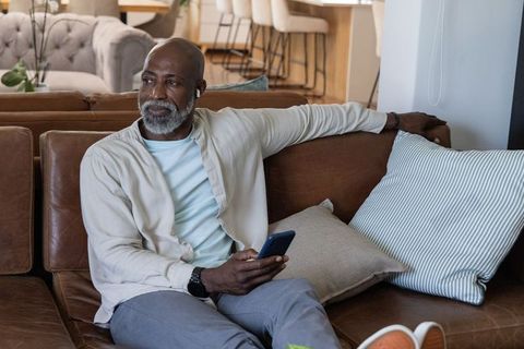 Senior african american man relaxing on leather sofa with smartphone and wireless earbuds