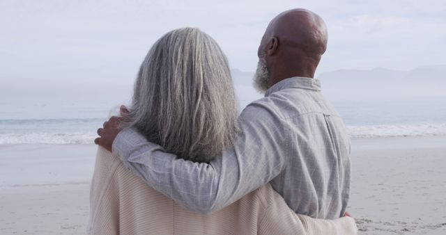 Senior couple standing arm in arm, gazing at calm ocean from sandy beach, evoking feelings of love and companionship. Useful for themes like senior living, retirement, romance, caregiving, and life enjoyment. Perfect for ads, blogs, and publications dedicated to relationships, elderly well-being, and serene lifestyles.