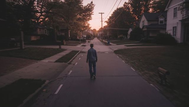 Solitary man walking down tree-lined suburban street at dusk with warm sunset silhouette calm mood