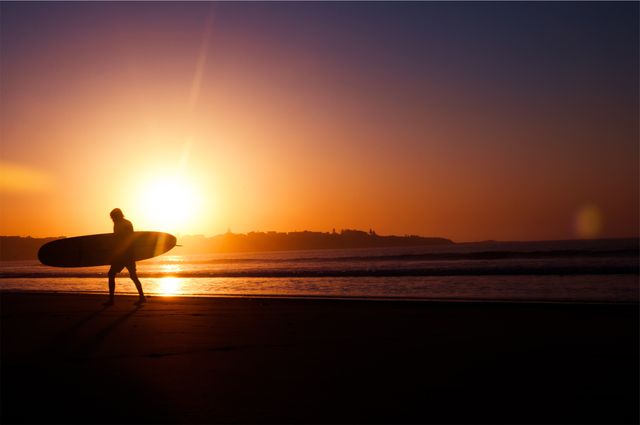 Silhouette of a surfer carrying a surfboard while walking on the beach during sunset. Ideal for use in travel, adventure, leisure, and lifestyle content. Captures the essence of beach culture, surfing enthusiasm, and tranquil sunset moments.