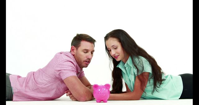 Young Caucasian couple lying down on floor discussing savings with pink piggy bank. Great for themes related to financial planning, budgeting, money management, teamwork, savings, and young couples building future together.