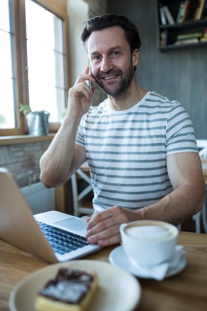 Portrait of smiling man talking on the mobile phone and using laptop in coffee shop