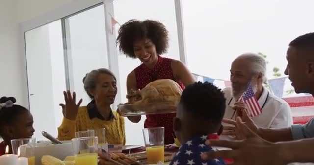 Scene captures an African American multi-generation family gathered around dinner table, celebrating Independence Day. A joyful moment as mother places turkey at the center of the festive meal. Table adorned with small US flags symbolizing patriotism. Perfect for illustrating joyful family gatherings, Independence Day celebrations, or highlighting American cultural traditions.