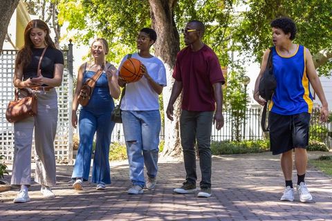 Diverse friends walking through sunlit neighborhood carrying basketball and backpacks