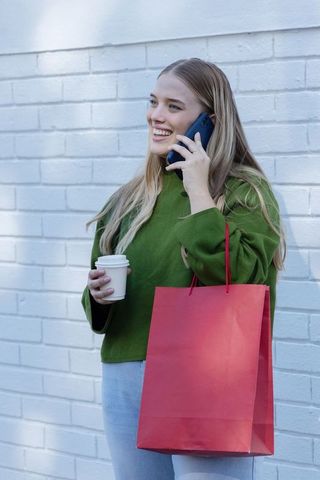 Woman talking on smartphone holding red shopping bag and coffee by white brick wall