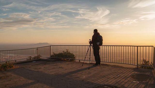 Landscape photographer standing at overlook adjusting tripod camera during golden sunset