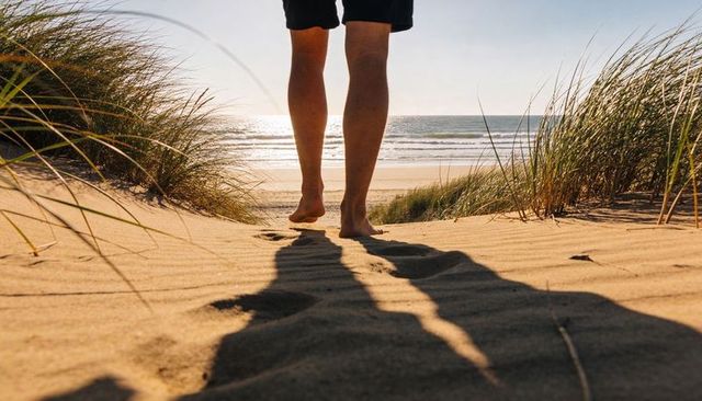 Barefoot walking on sunlit dune path toward ocean horizon with footprints and beach grass