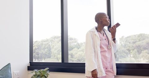 Clinician recording voice note on smartphone while looking out window in medical office