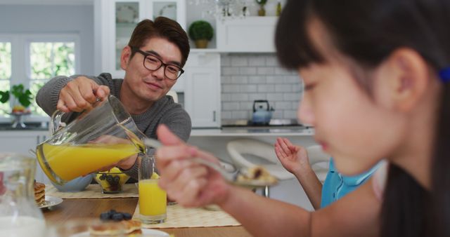 Happy asian father in kitchen having breakfast pouring juice for smiling son and daughter. happy family, at home in isolation during quarantine lockdown.