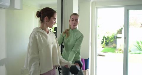 Female friends training with dumbbell in sunlit living room offering personal coaching