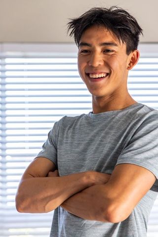 Confident young asian man smiling with arms crossed by window blinds in grey tee