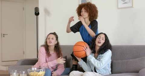 Friends reacting to basketball game on sofa holding ball and snacks, casual indoor
