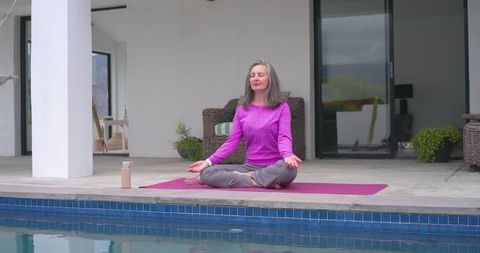 Senior woman meditating poolside on magenta mat wearing purple top and gray pants, calm