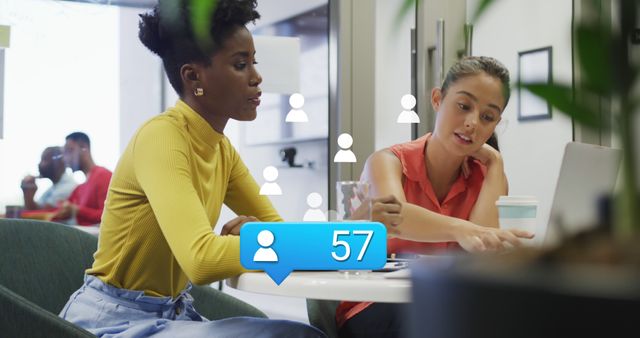 Two women engaged in a discussion while working on a laptop in a modern office setting. Visible social media icon illustrates focus on digital and social media work. Ideal for use in articles, blogs, and advertisements related to professional collaboration, social media strategies, and modern work environments.