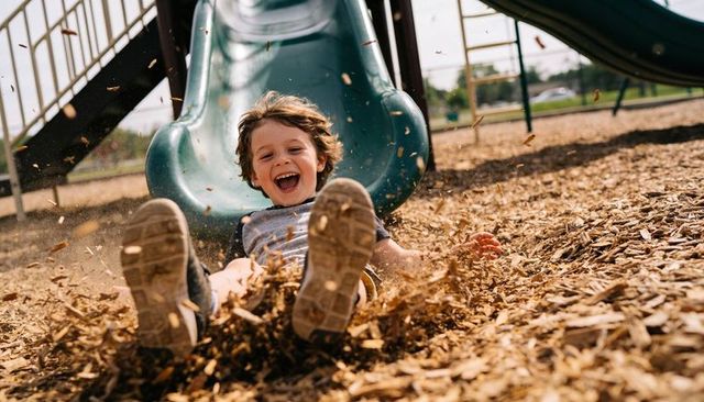 Joyful young boy sliding into wood chips at playground, sneakers kicking, summer fun