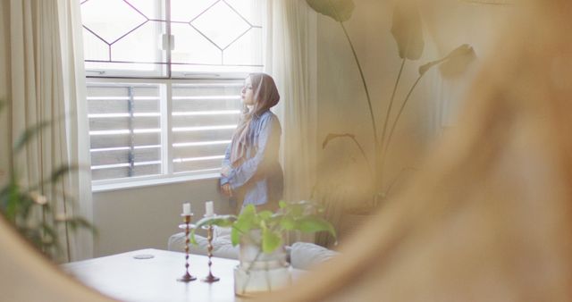 Woman in headscarf gazing out bright window in serene living room - Download Free Stock Images Pikwizard.com