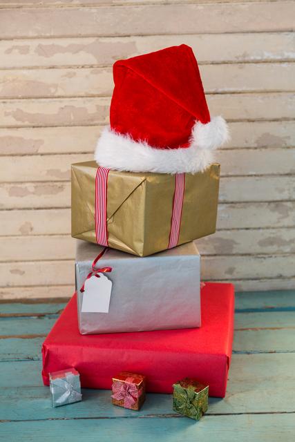 Stacked Christmas gifts wrapped in red, gold, and silver paper with a Santa hat on top, placed on a wooden plank. Ideal for holiday greeting cards, festive advertisements, and Christmas-themed promotions.