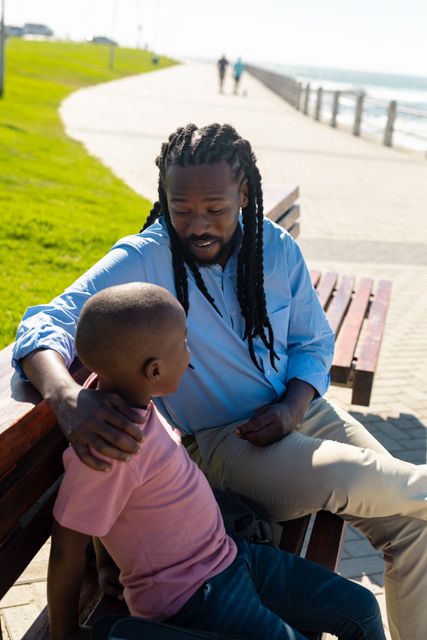 African american father with braided hair talking to son while sitting on bench at promenade. unaltered, parenting, family, lifestyle and togetherness concept.