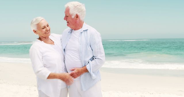 Senior couple enjoying each other's company on a beautiful, sunny beach. Both are smiling and dressed in light clothing, suggesting a relaxed and happy mood. Ideal for use in advertisements for retirement, vacation packages, health and wellness industries, or articles focusing on love and companionship in later years.