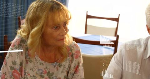 Senior woman sitting at dining table turning toward companion with data and chart overlays