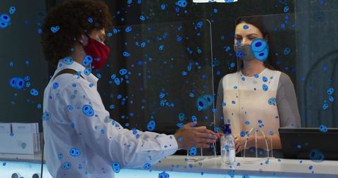 Receptionist greeting client behind plexiglass divider wearing masks while using sanitizer