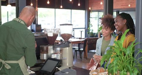 Smiling friends ordering coffee at counter in sunlit cozy modern cafe with pastry display