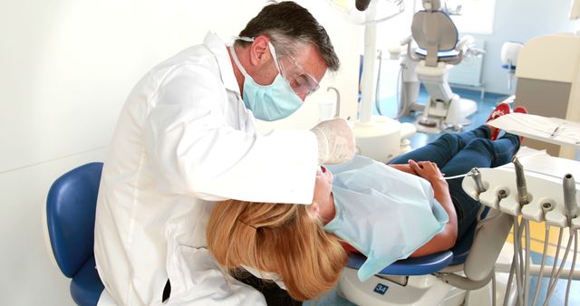Image depicts a dentist in protective gear treating a female patient. Useful for medical and dental themes, healthcare services, patient care, dental health articles, and promoting professional dental practices. Highlights the importance of regular dental check-ups and modern dental care environments.