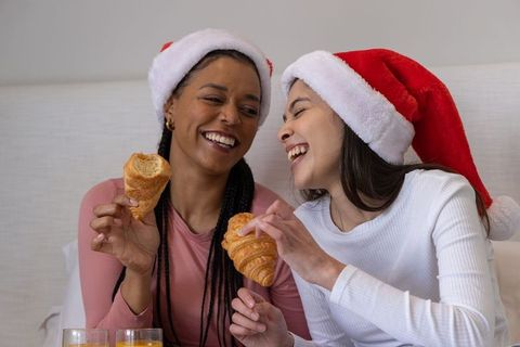 Two women wearing Santa hats laughing and sharing croissants during cozy holiday morning