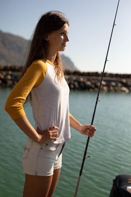 Teenage Caucasian girl, enjoying her time on a promenade by the sea, on a sunny day, holding a fishing rod, looking away. 
