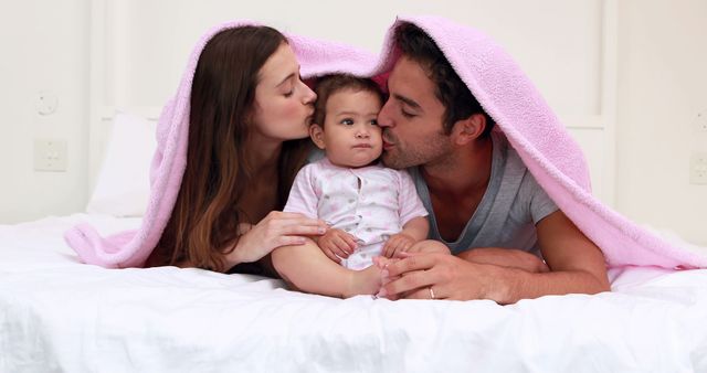 Loving parents kissing their baby on the bed under pink blanket