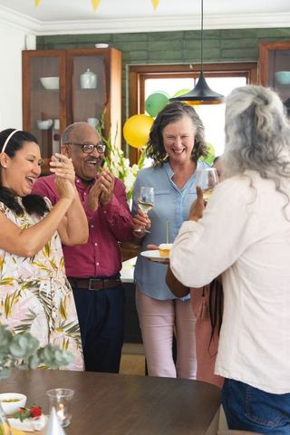 Mature friends celebrating birthday in home kitchen, clapping while host presents cake