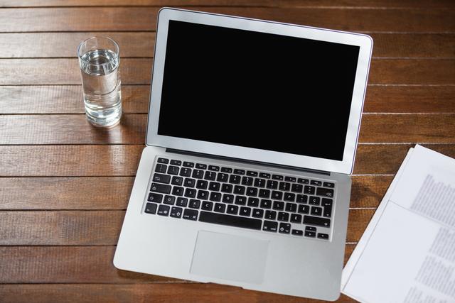 This image shows a laptop placed on a wooden table, accompanied by a glass of water and some documents. This scene is ideal for illustrating workspaces, business environments, or technology use in professional settings. It can be used for blog posts, office-related articles, presentations, or marketing materials focusing on productivity and modern work setups.
