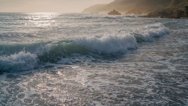 Breaking ocean waves rolling toward sandy shore at hazy sunset with rocky headlands